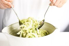 a person is holding two spoons over a bowl full of shredded green vegetables on a white table