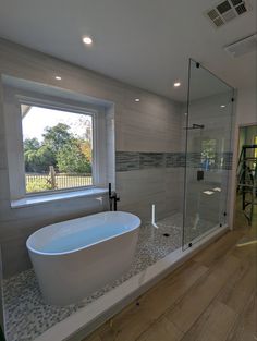 a bath tub sitting next to a large window in a bathroom with wooden floors and white walls