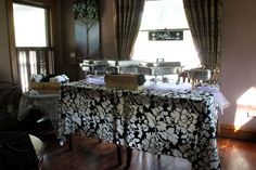 a dining room table covered with a black and white damask cloth next to two windows