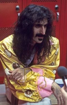 a man with long hair sitting in front of microphones and wearing a yellow robe