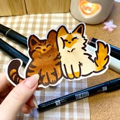 a person holding up a sticker with two cats on it next to some pens