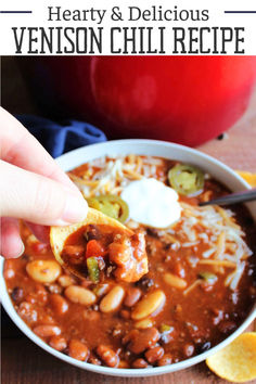 a hand holding a tortilla chip over a bowl of hearty and delicious venison chili recipe