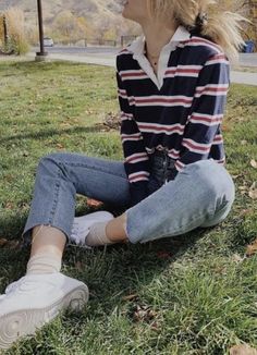 a woman sitting on the grass with her legs crossed wearing jeans and a striped shirt