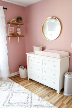 a baby's room with pink walls, white furniture and a round mirror on the wall