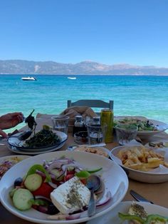 a table with plates and bowls of food on it next to the ocean in front of some mountains