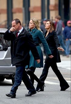 three people walking down the street in front of a car, one man is holding his hand up to his ear