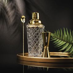 a glass bottle and two golden spoons sitting on a tray next to a fern