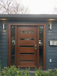 a wooden door is shown in front of a gray house with plants and bushes around it