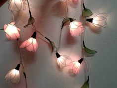 pink flowers with green leaves are hanging on the wall next to a white wall and some string lights