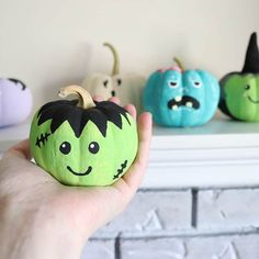 a hand holding a green pumpkin with faces painted on it's face and three other small pumpkins in the background