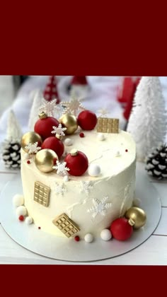 a white cake with red and gold decorations on it sitting on a table next to pine cones