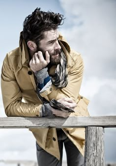 a man leaning on a wooden fence talking on his cell phone while wearing a trench coat