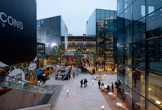 people are walking around an outdoor shopping mall at night, with lights on the walkway and large glass buildings in the background