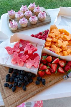 various fruits and cupcakes are on a cutting board next to a tray with muffins, strawberries, and watermelon