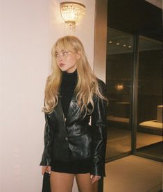 a woman standing in front of a white wall wearing a black jacket and skirt with high heels