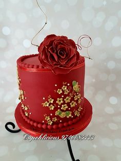 a red cake with gold decorations and a rose on top that is sitting on a metal stand