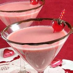 two martini glasses filled with pink liquid and topped with cherries on a red tablecloth