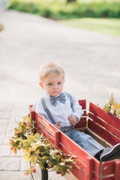 a little boy in a bow tie sitting in a red wagon with leaves on it