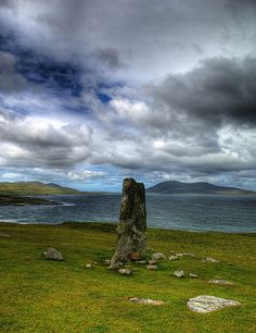 a large rock sitting on top of a lush green field next to the ocean under a cloudy sky