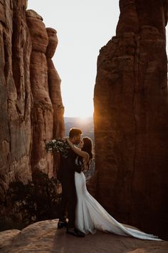 a bride and groom standing on top of a rock formation with the sun behind them