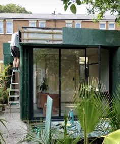 a man standing on a ladder in front of a green building next to trees and plants