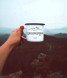 a hand holding a coffee cup with the words groomsman on it and mountains in the background