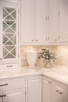 a white kitchen with marble counter tops and cabinets in the corner, vase on top