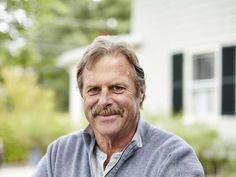 a man with a moustache is smiling at the camera while standing in front of a house