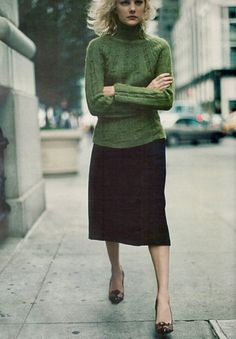 a woman in green sweater and black skirt walking down the street with her arms crossed