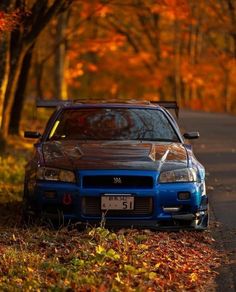 a blue car parked on the side of a road in front of trees with orange leaves