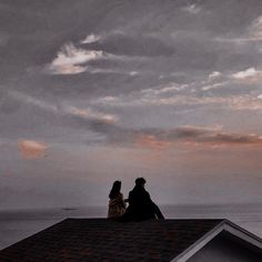 two people sitting on top of a roof looking out at the ocean and clouds in the sky