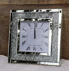 a square clock with roman numerals on the face is sitting in front of a stone wall