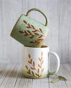 two coffee mugs sitting on top of each other, one with a handle and the other with leaves painted on it