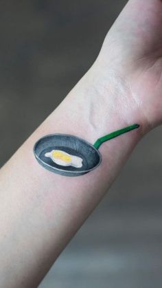 a person with a small tattoo on their arm holding an egg in a frying pan