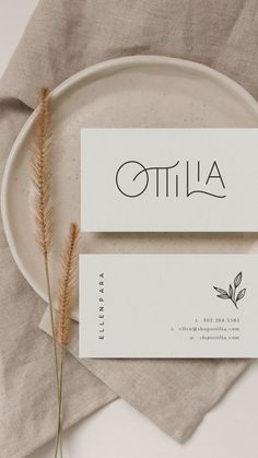 two business cards sitting on top of a white plate next to some dried plants and a napkin