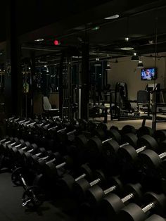 an empty gym with rows of black dumbbells in the foreground and a red light at the far end