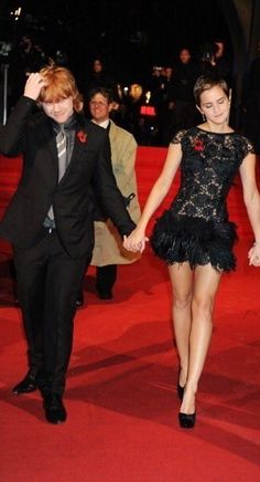 two people holding hands while walking down a red carpeted area with other people in the background