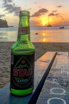 a bottle of beer sitting on top of a wooden table next to the ocean at sunset