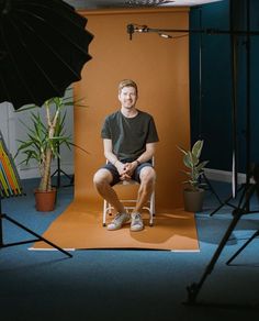 a man sitting on a chair in front of an orange backdrop with two plants behind him