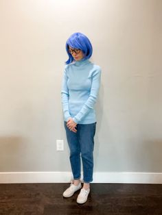 a woman with blue hair standing in front of a white wall wearing jeans and a light blue sweater