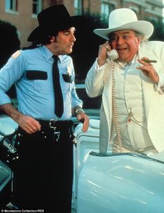 two men standing next to each other in front of a car talking on the phone