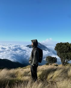 a man standing on top of a grass covered hill next to a forest filled with clouds