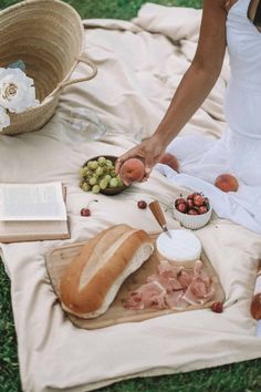 a woman in white dress sitting on blanket next to bread and grapes with other food items