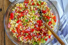 a glass bowl filled with corn, tomatoes and green onions next to a wooden spoon