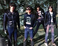 four young men standing next to each other in front of some trees and bushes, wearing black leather jackets