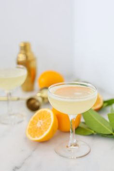 two glasses filled with white wine next to oranges on a marble counter top and green leaves