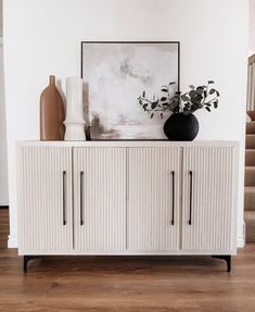 a white cabinet with two vases and a painting on the wall next to it