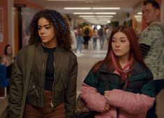 two young women walking down a hallway with other people in the background and onlookers