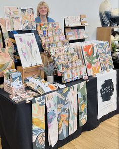 a woman standing next to a table with paintings on it and other items for sale