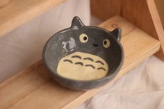 a black and white ceramic bowl with a totoro face on the front, sitting on a wooden shelf